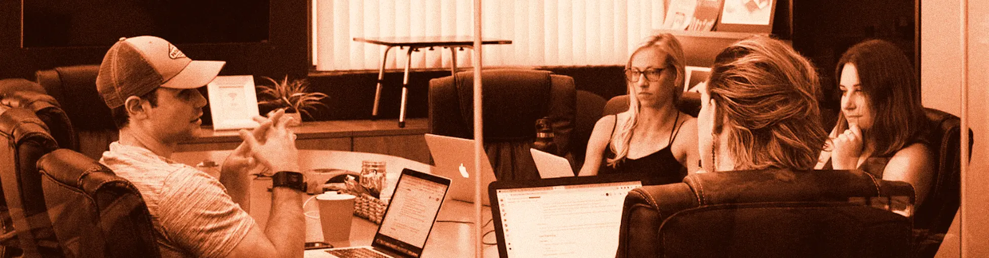 Four colleagues around a conference table with laptops, one speaking and the others listening.