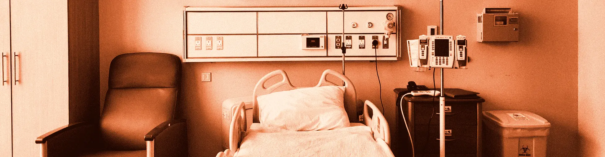 A hospital room with an empty bed, a headwall panel, an infusion pump on a stand, a red recliner and a yellow biohazard bin.