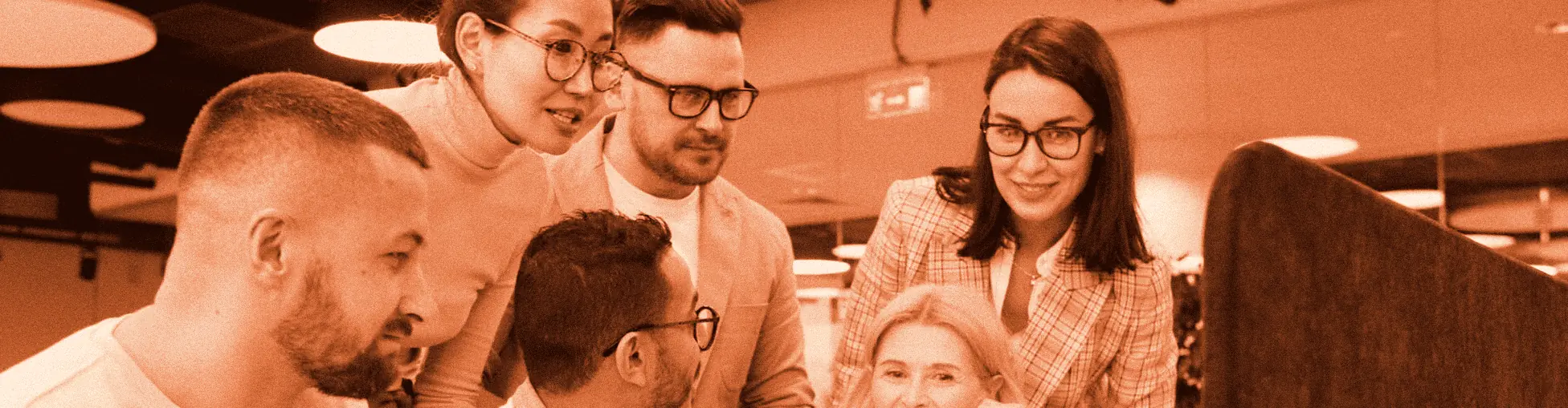 Five colleagues gathered around a laptop, one pointing at the screen while the others lean in and smile.