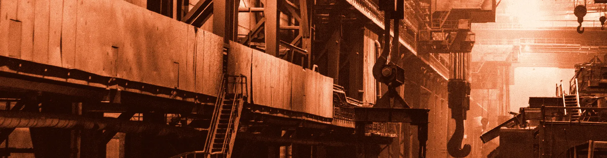 Inside a steel plant: overhead cranes, a large lifting hook, walkways and steel frames lit in amber.